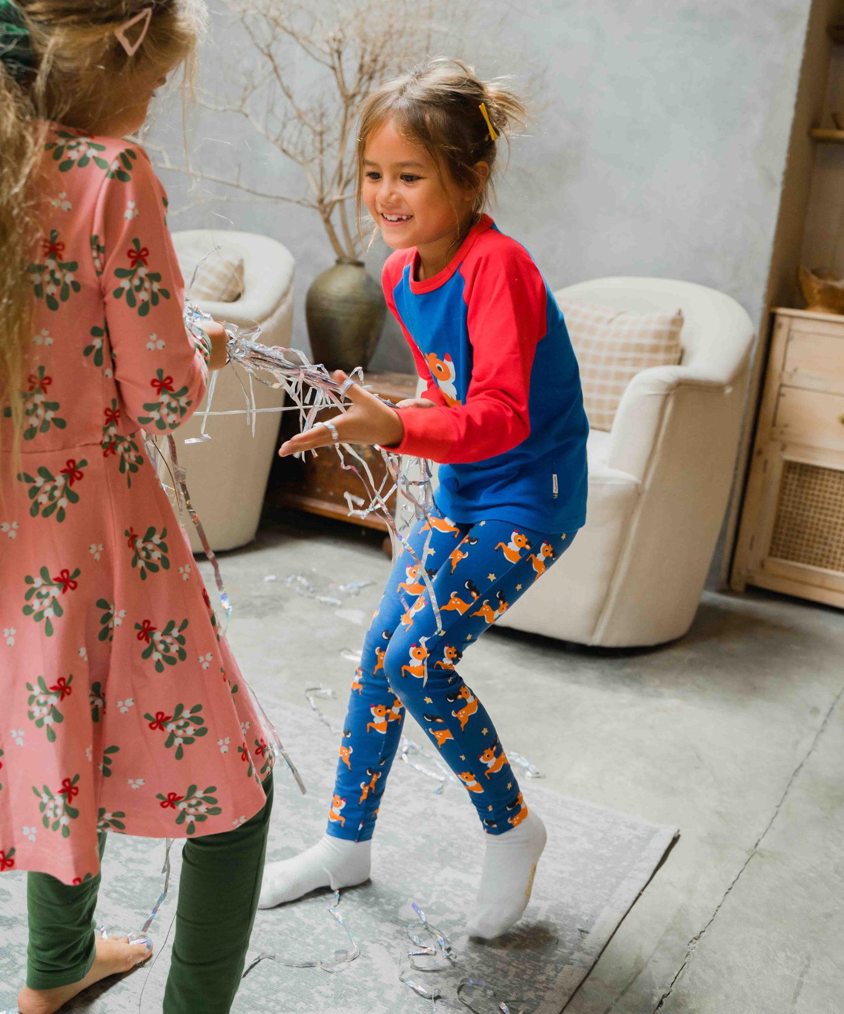 A child wearing the Maxomorra Reindeer Leggings with a coordinating reindeer print raglan top. The child is playing with Christmas streamer decorations. 