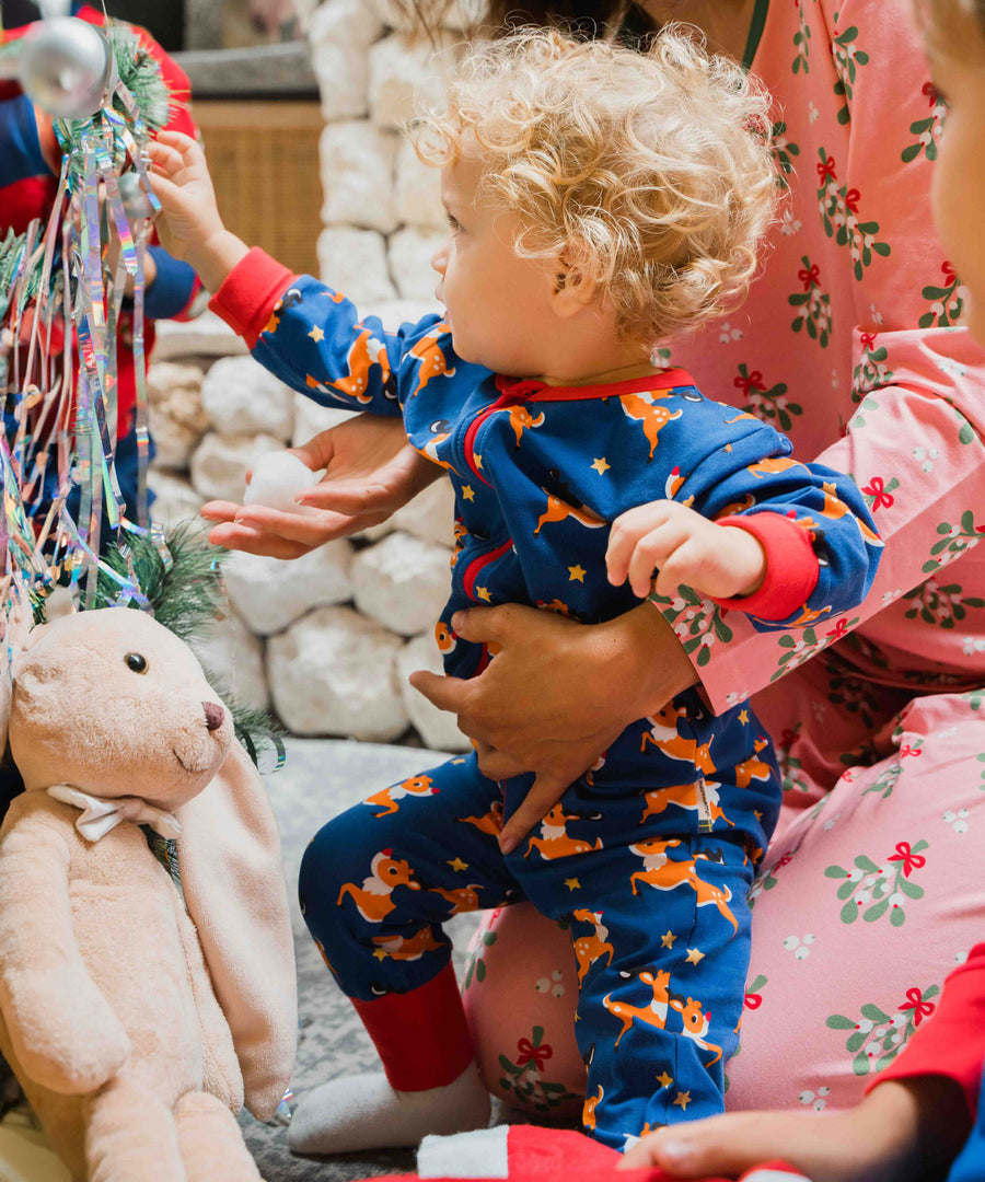 A young child wearing the Maxomorra Reindeer Long Sleeve Rompersuit. The child is reaching for a bauble from the Christmas tree. 