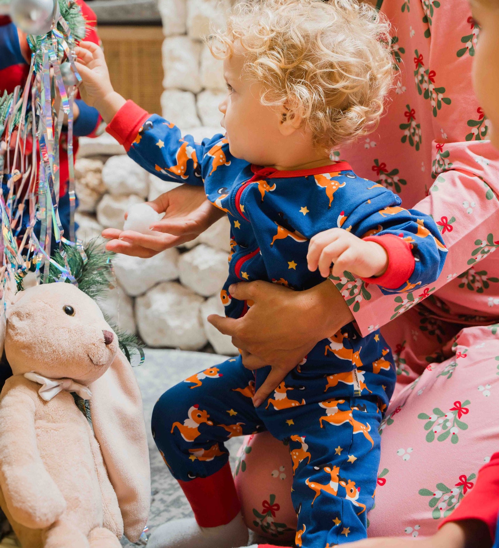 A young child wearing the Maxomorra Reindeer Long Sleeve Rompersuit. The child is reaching for a bauble from the Christmas tree. 