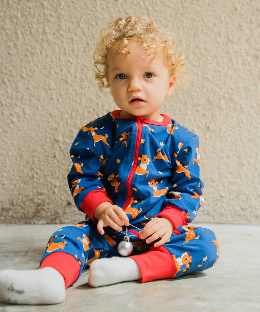 A young child wearing the Maxomorra Reindeer Long Sleeve Rompersuit. The child is sitting in front of a beige coloured wall. 