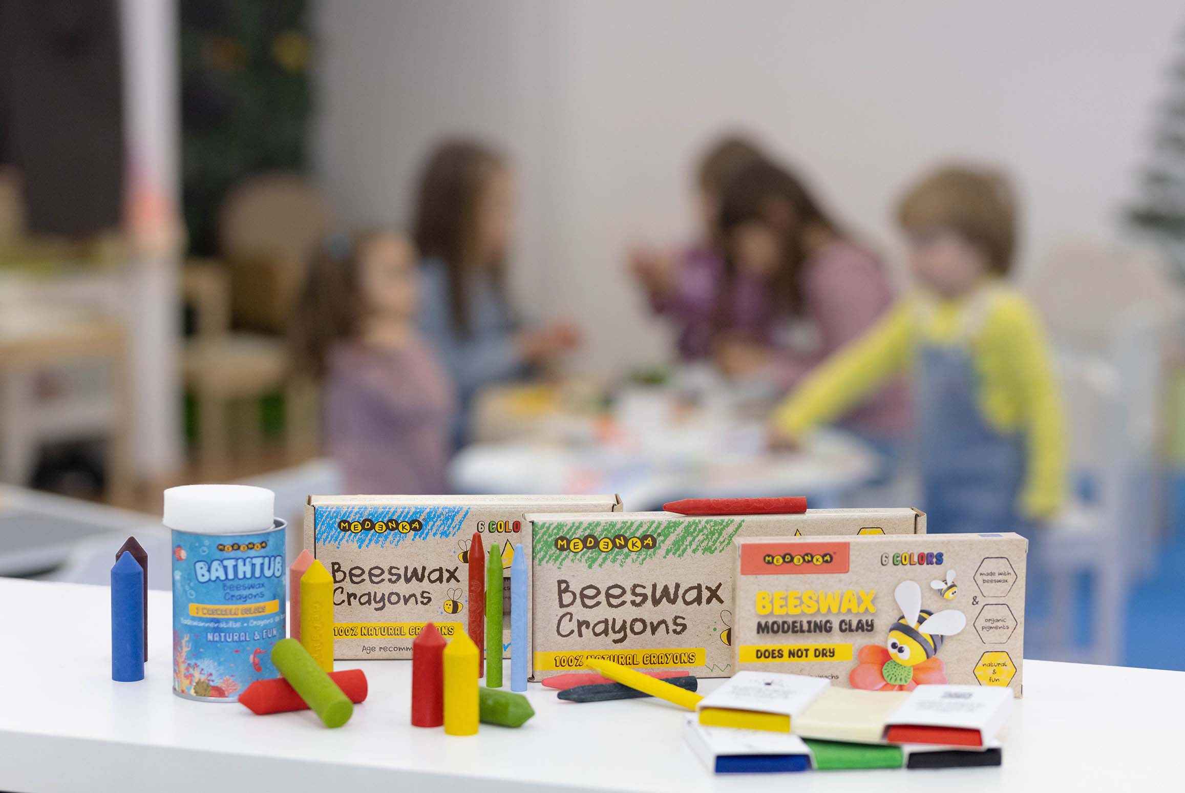 A display of the Medenka beeswax crayon and modelling clay range, with children colouring in the background. 