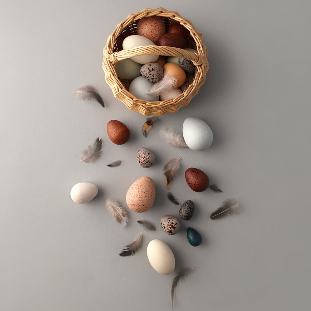 A brown floor with the Moon Picnic A Dozen Bird Eggs in a Basket set. 7 different sized and coloured eggs lay on the floor next to the basket with 4 fluffy feathers surrounding