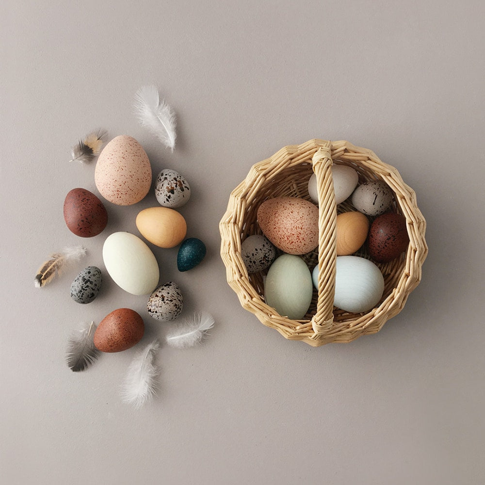 A grey floor with the Moon Picnic A Dozen Bird Eggs in a Basket set. 9 different sized and coloured eggs lay on the floor to the left of the basket with 5 fluffy feathers surrounding