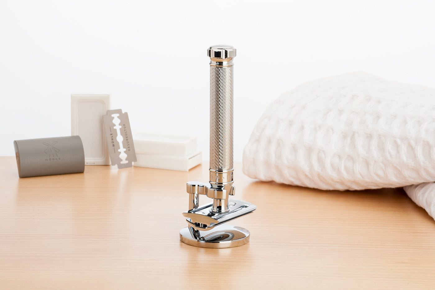 Close up of a Muhle stainless steel traditional safety razor in a chrome razor stand next to some spare blades and white towel