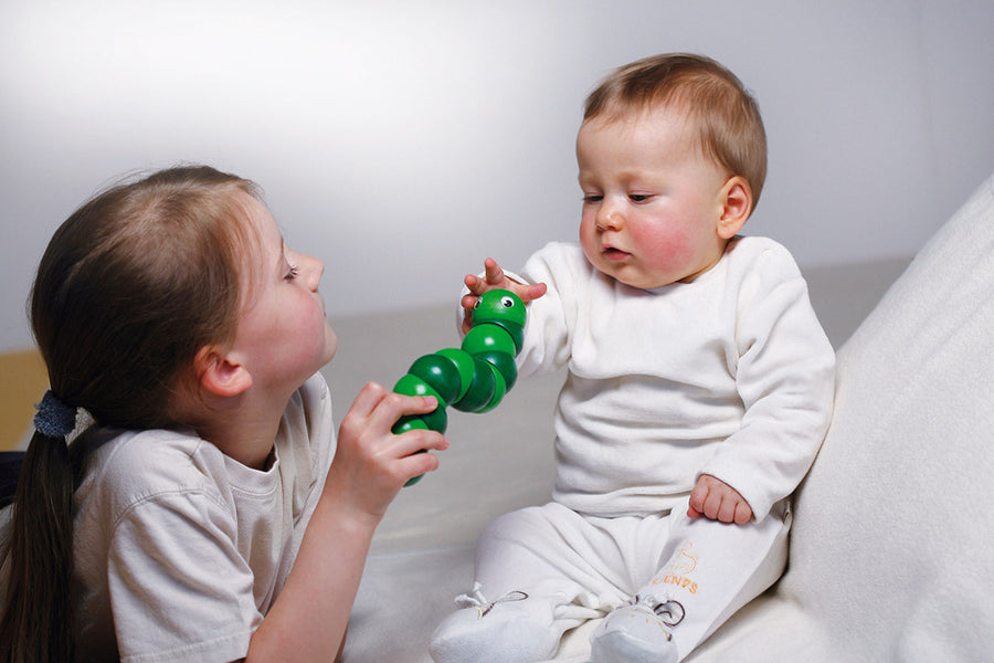 Young girl playing with a green Naef Juba worm toy in front of a baby who is reaching out to hold it.