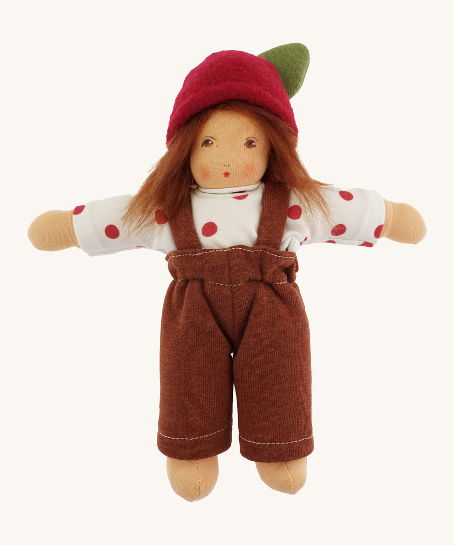 A beautiful Waldorf, handmade doll with a white top with red spots, a brown pair of dungarees, light brown hair and a red apple hat with a green leaf. On a cream background