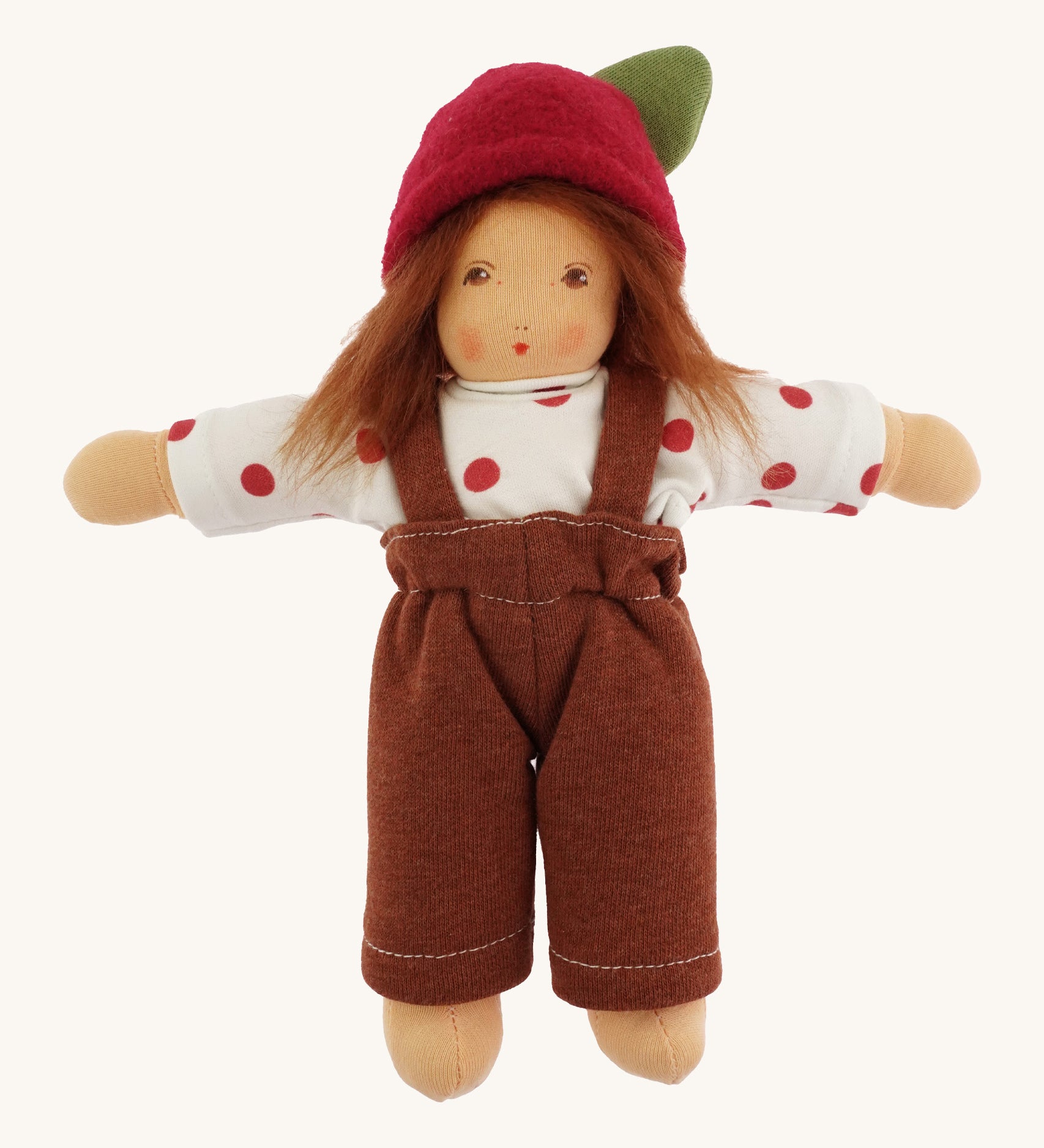 A beautiful Waldorf, handmade doll with a white top with red spots, a brown pair of dungarees, light brown hair and a red apple hat with a green leaf. On a cream background