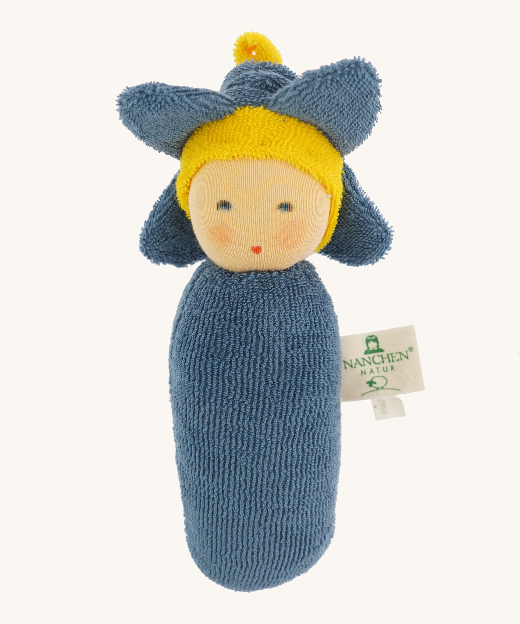 Nanchen Flower Rattle Doll - Forget-Me-Not, is a soft, organic and handmade sensory doll toy with a blue body, a little blue hat with blue forget-me-not leaves and a yellow strip of fabric to mimic hair. This doll also has an adorable face with blue eyes