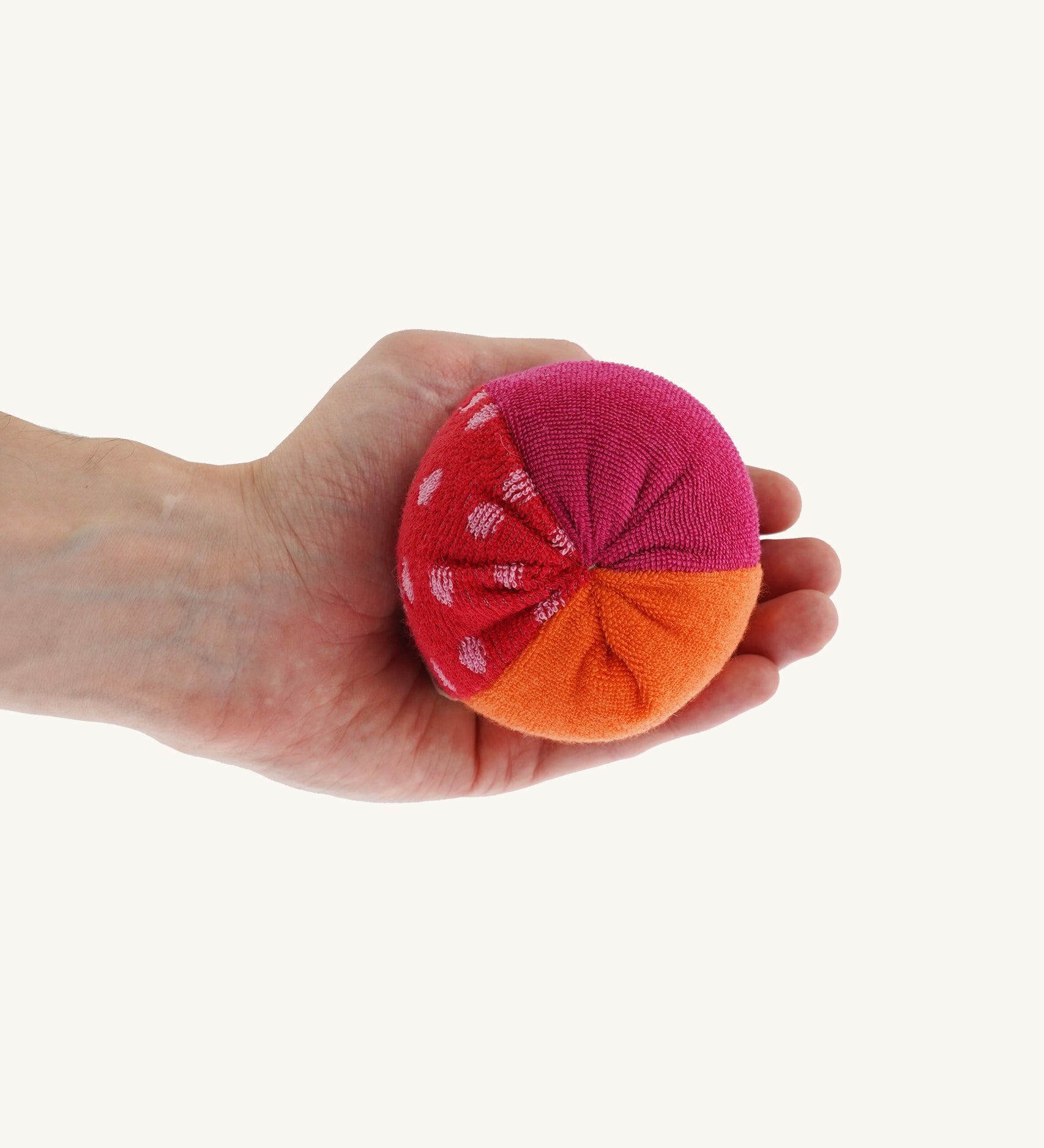 Nanchen Large Colourful, Organic Baby Rattle Ball on a hand for scale and colour. This is a soft terry material in red, orange and pink colours on a cream background.