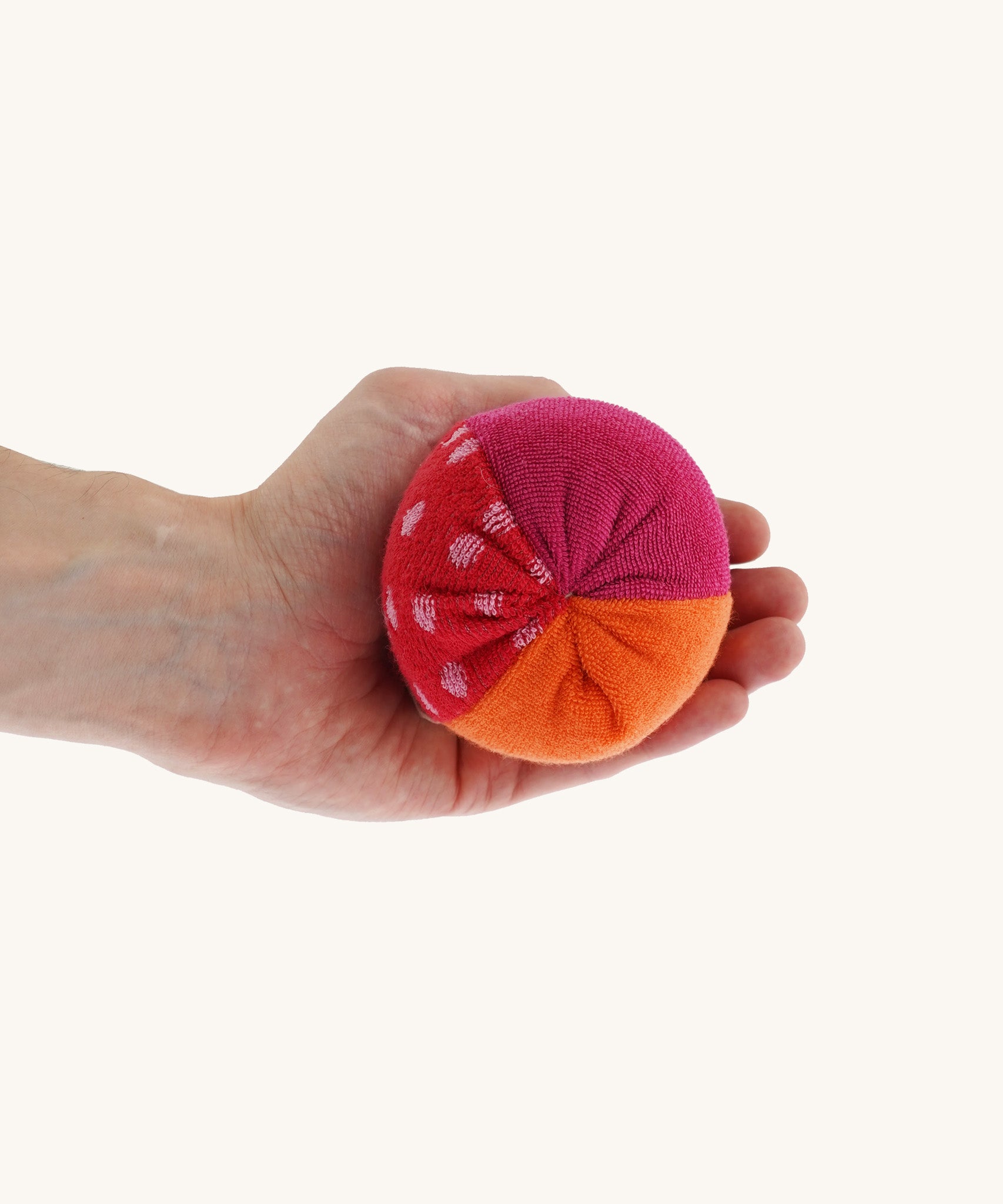 Nanchen Large Colourful, Organic Baby Rattle Ball on a hand for scale and colour. This is a soft terry material in red, orange and pink colours on a cream background.