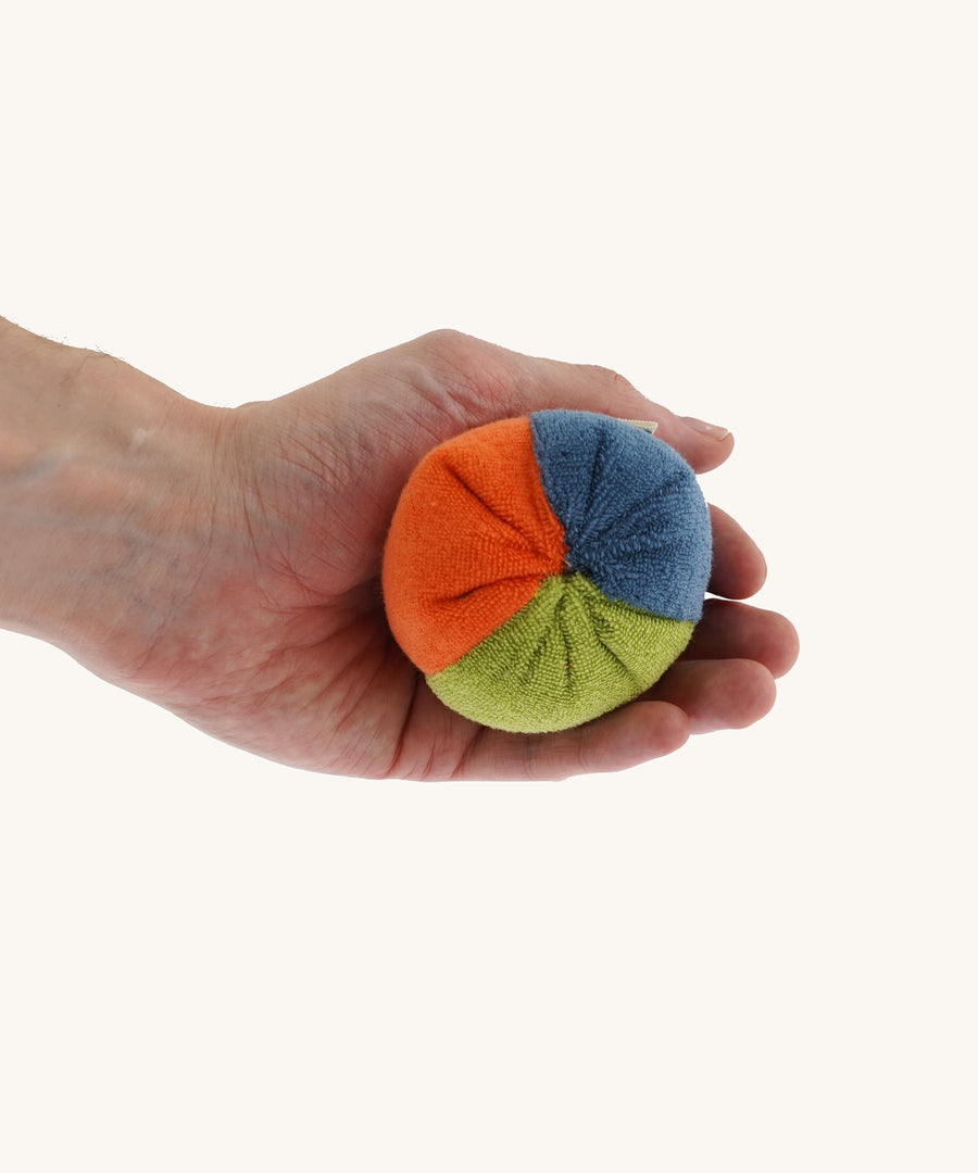 Nanchen small Colourful, Organic Baby Rattle Ball on a hand for scale and showing the different colours. This is a soft terry material in blue, orange and green colours on a cream background.