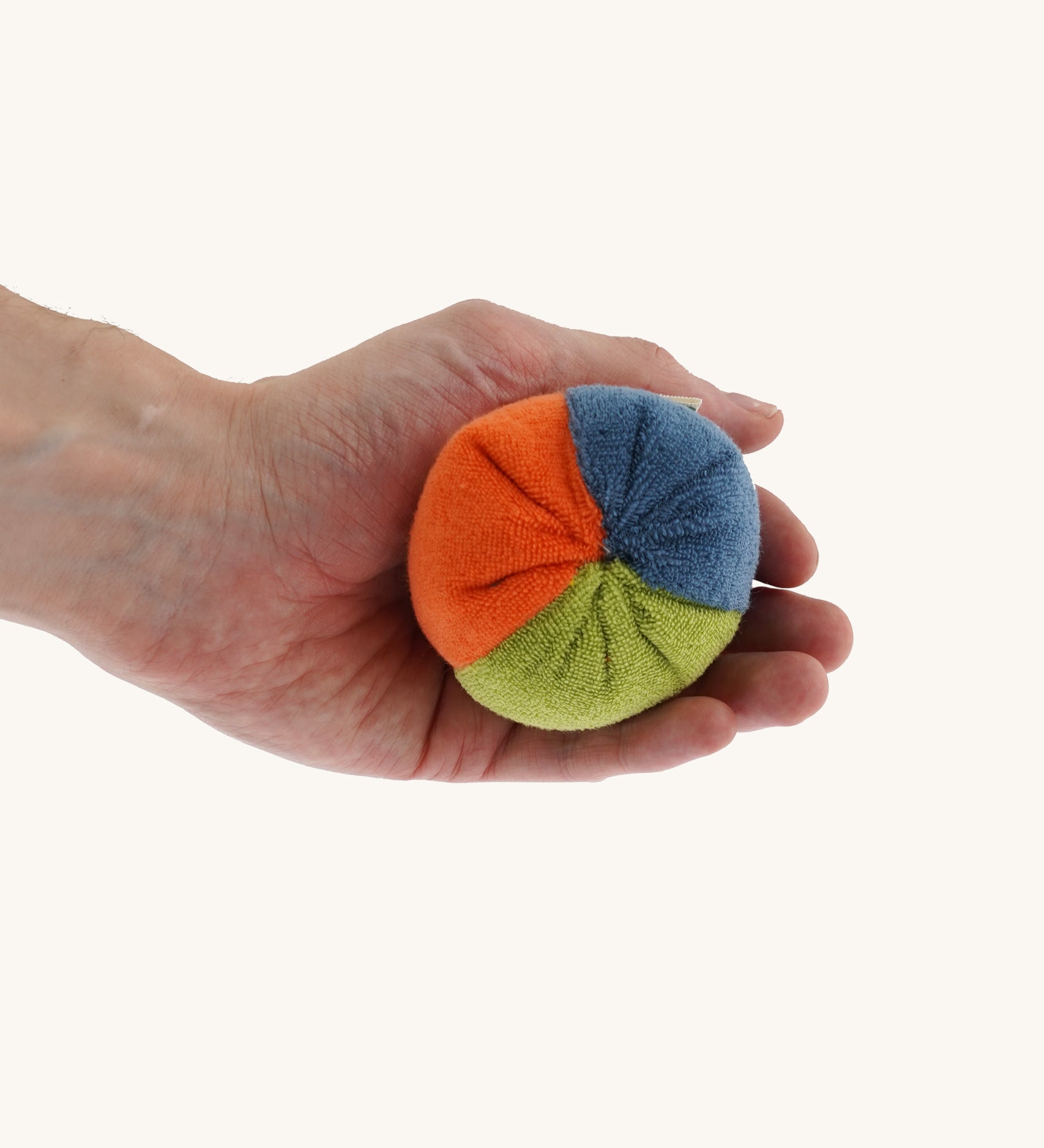 Nanchen small Colourful, Organic Baby Rattle Ball on a hand for scale and showing the different colours. This is a soft terry material in blue, orange and green colours on a cream background.