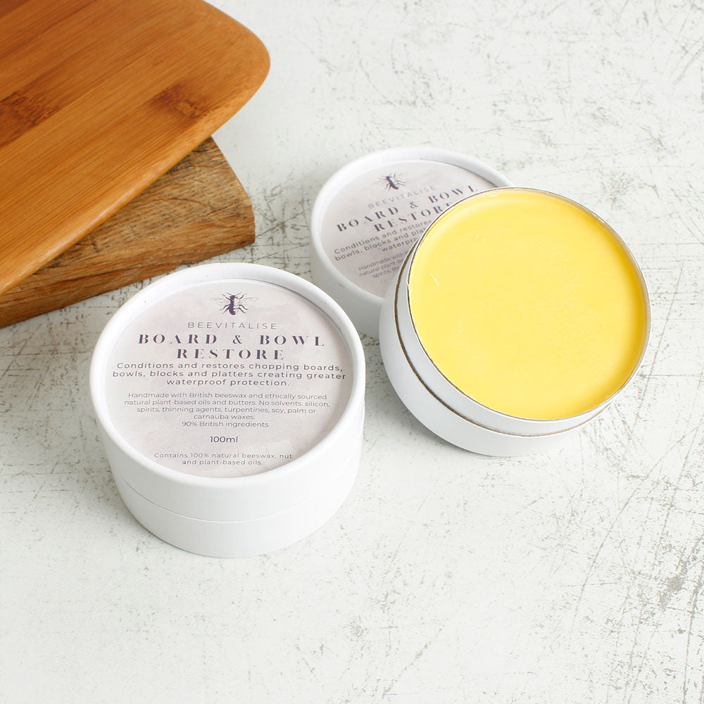 Two tubs of Oakdale Bees Beevitalise natural beeswax wooden board and bowl balm on a wooden floor next to a wooden chopping board