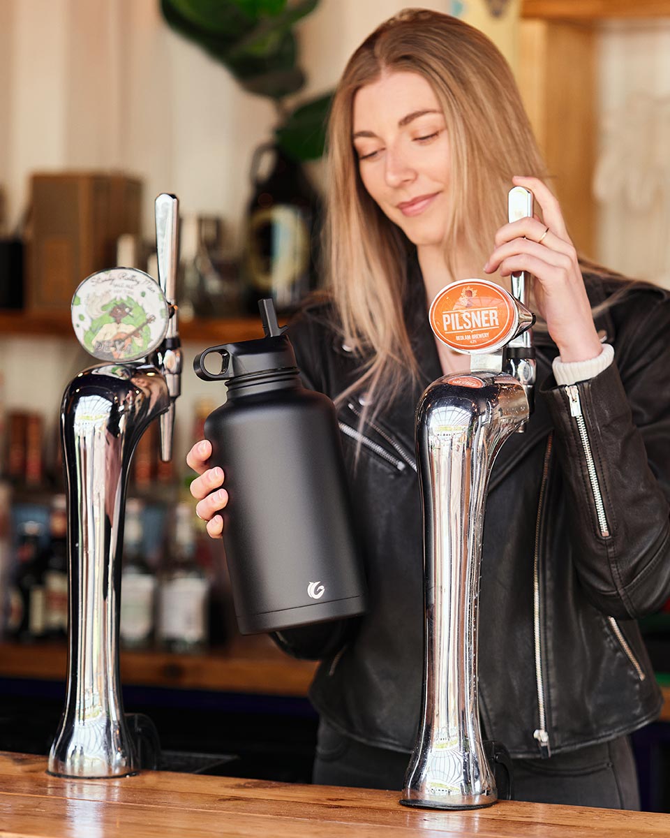 Woman holding a One Green Bottle plastic free stainless steel water bottle next to some beer pumps 