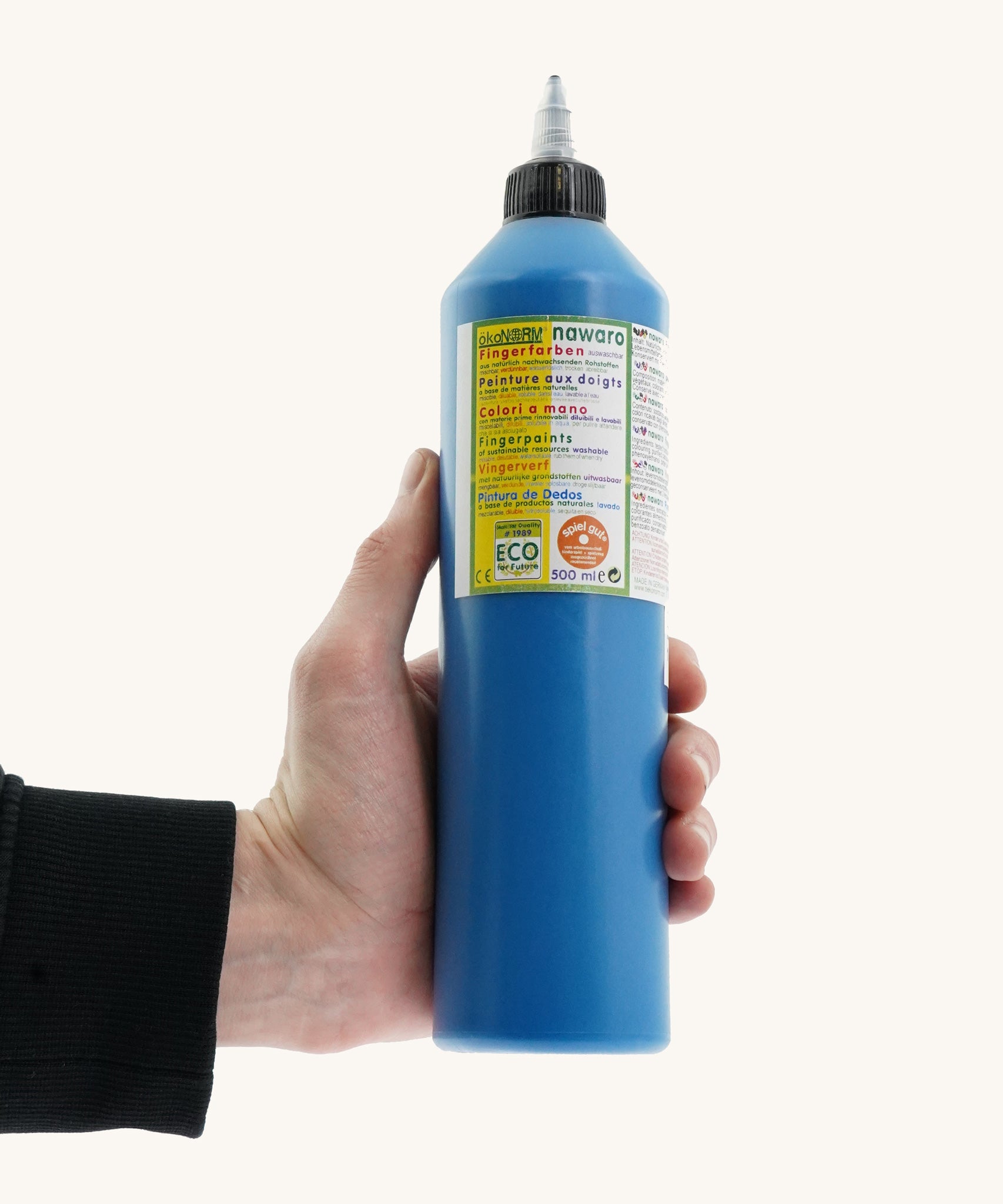 An adult's hand holding a 500ml bottle of OkoNorm finger paints in a blue colour. 