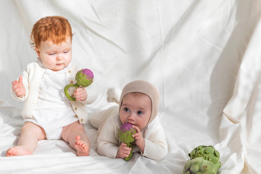 One baby sitting holding an Oli & Carol Artichoke Rattle Teether toy with another baby lying by their side chewing on an Oli & Carol Artichoke Rattle Teether