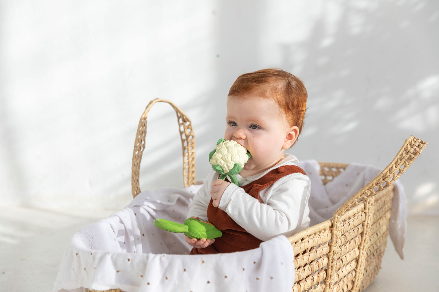 A baby sitting up in a moses back and chewing on an Oli & Carol Cauliflower Rattle Teether with a corn teether in the other hand