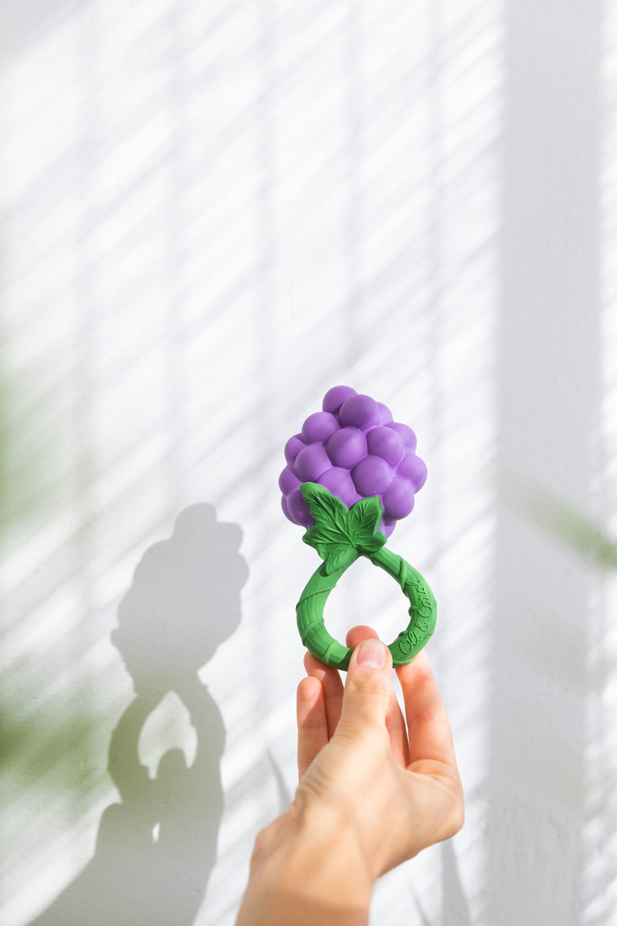 An adult's hand holding up the Oli & Carol Grape Rattle Teether