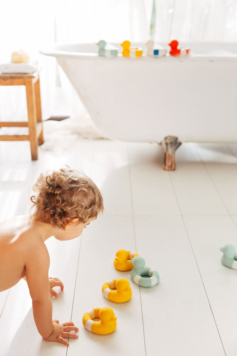 Child in bathroom playing with yellow and mint coloured Oli & Carol Flo The Floatie Ducks natural rubber bath toys