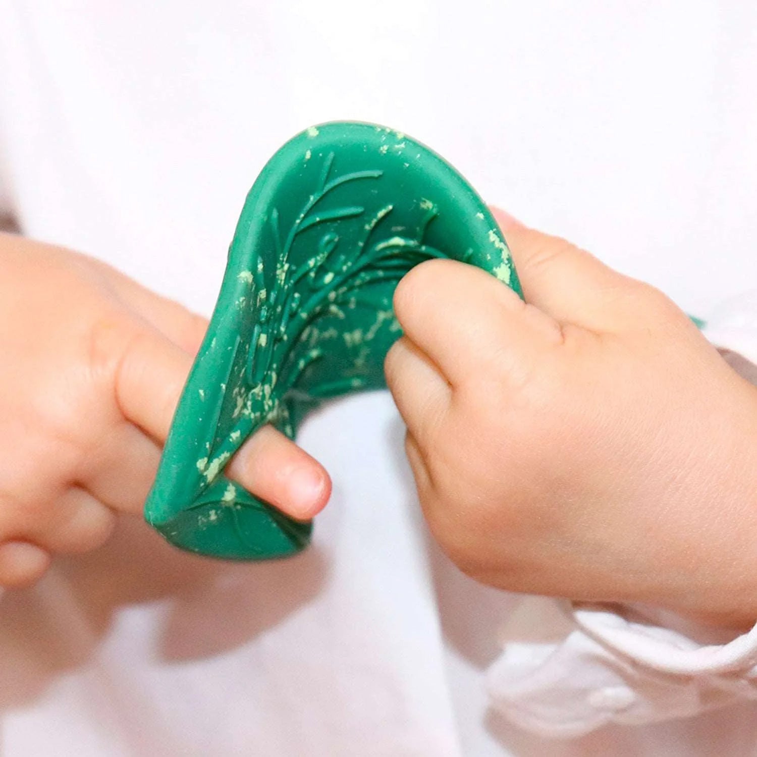 A close up of a baby holding onto the Oli & Carol green coloured Chlorophyll Leaf Baby Teether showing the flexibility