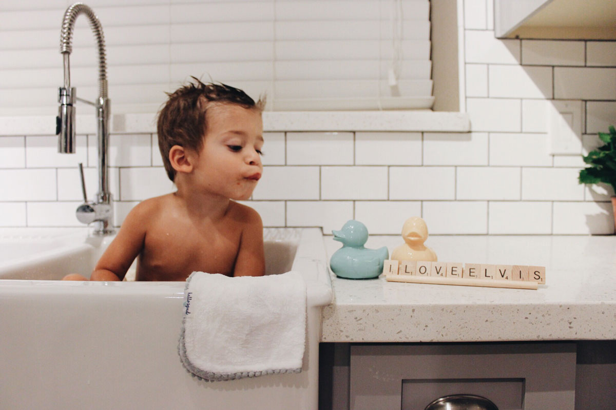 Child bathing in a kitchen sink with a mint and nude coloured Oli & Carol Elvis The Duck Bath Toys placed on the counter top by their side