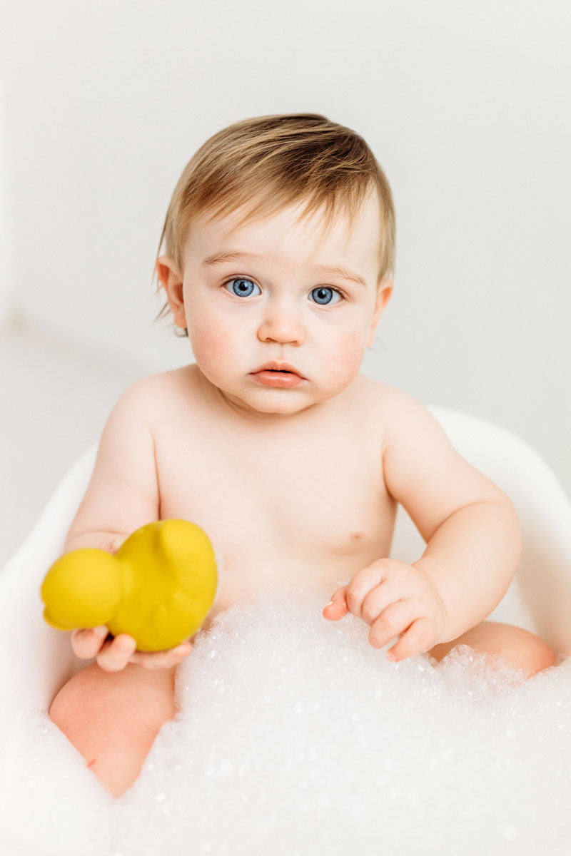 Baby in a small bath tub holding onto a yellow Oli & Carol Elvis The Duck Bath Toy