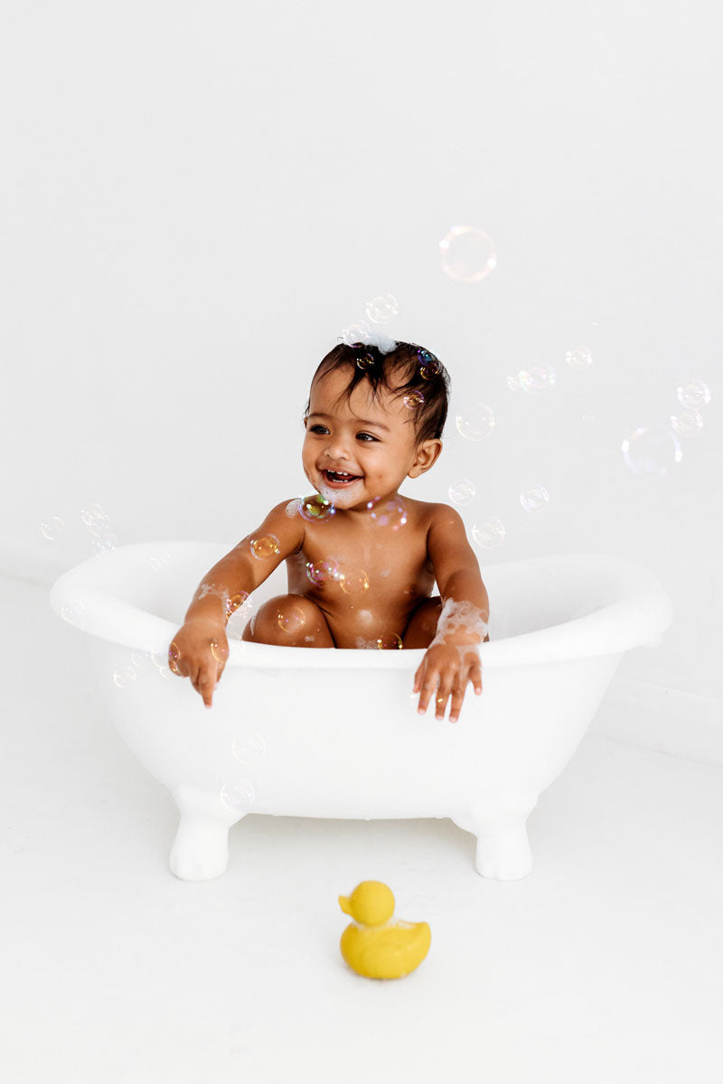 Baby in a small bath tub holding with bubbles floating around with a yellow Oli & Carol Elvis The Duck Bath Toy on the floor in front