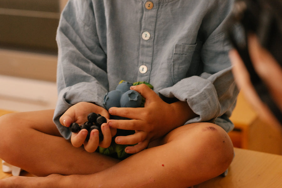 Close up a child holding a bunch of real blueberries next to the oli and carol eco-friendly natural rubber jerry blueberry toy