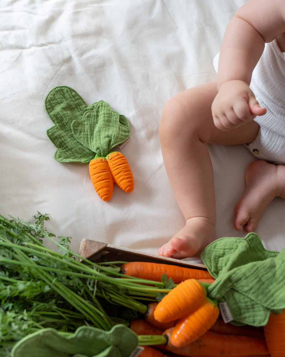 Oli & Carol natural rubber cathy the carrot mini baby teething toy on a white bed next to a crate of carrots and a baby's leg