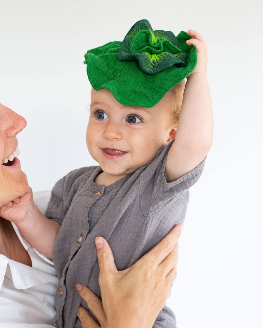 Woman holding a baby that is holding up the Oli & Carol mini kale baby comforter toy above its head