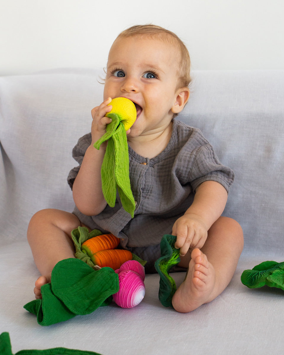 Baby sat on a grey sheet sucking on the Oli & Carol eco-friendly mini lemon baby teething toy