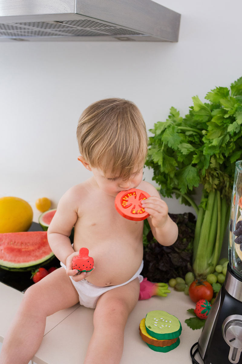 Child chewing on teh Oli & Carol Renato The Tomato with a pile of other Oli & Carol toys by their side