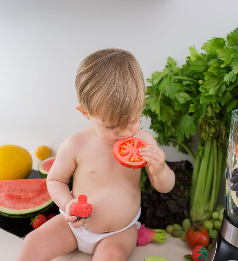 Child chewing on teh Oli & Carol Renato The Tomato with a pile of other Oli & Carol toys by their side