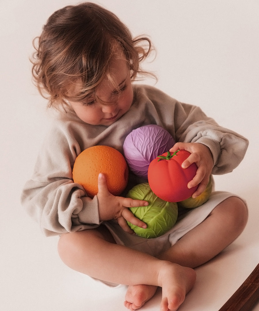 Toddler with an arm full of different fruit and vegetable Oli & Carol 100% Natural Rubber Baby Sensory Balls