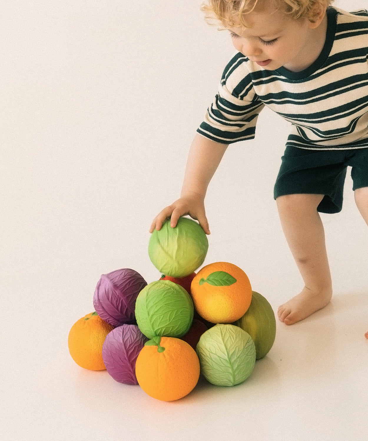 Toddler with a pile of different fruit and vegetable Oli & Carol 100% Natural Rubber Baby Sensory Balls