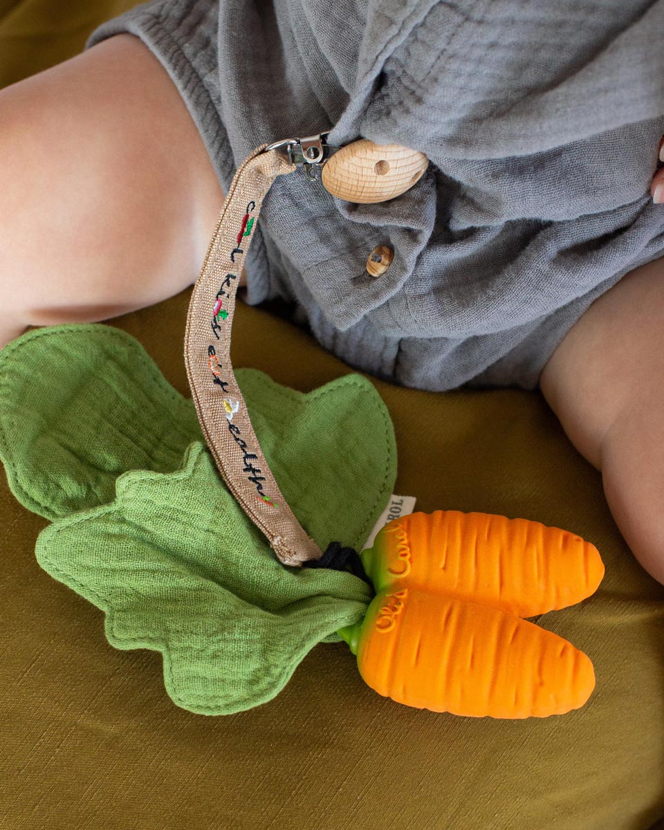 Close up of the Oli & Carol natural rubber carrot baby teething toy on a green sheet in front of a baby