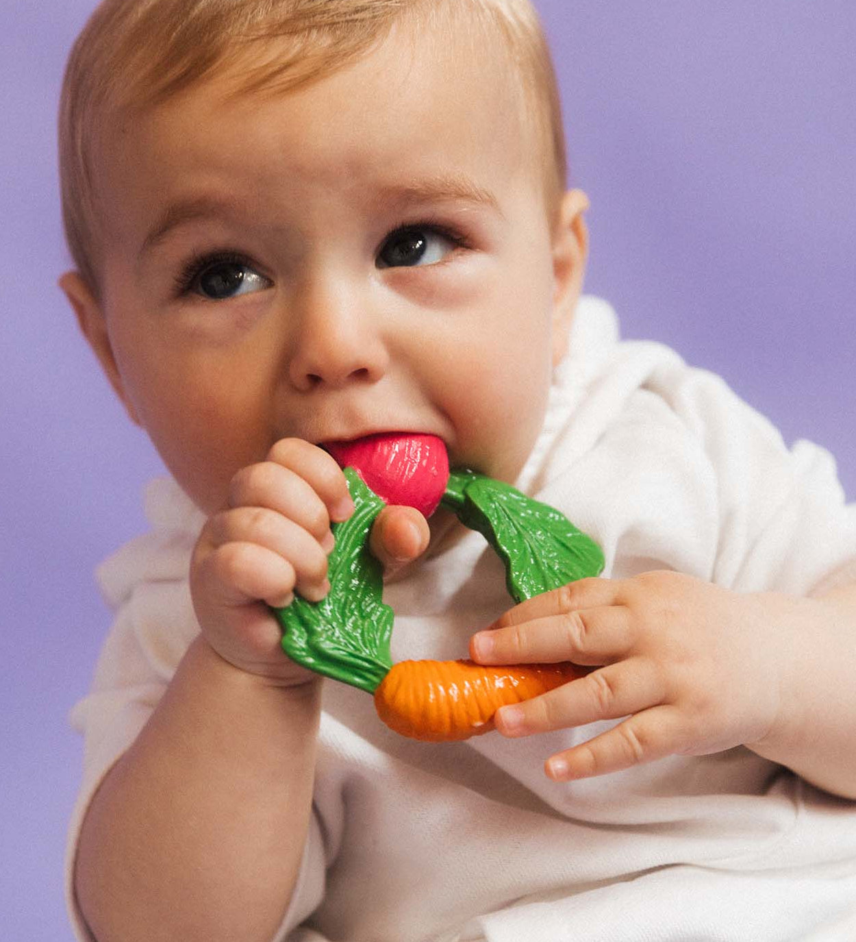 A baby clutching the Oli & Carol Natural Rubber Veggie Teething Ring.