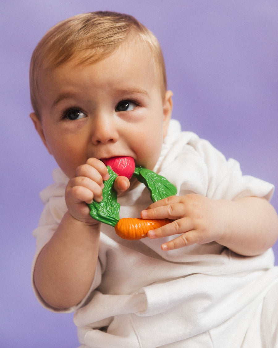 A toddler sits with four Oli & Carol 100% Natural Rubber Baby Teething Rings at their feet