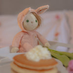 Olli Ella pink coloured Babbit Bunny Dinky Dinkum Doll in a seated position on a blanket. A pile of pancakes can be seen at the front. 