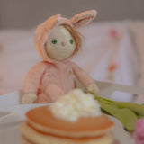 Olli Ella pink coloured Babbit Bunny Dinky Dinkum Doll in a seated position on a blanket. A pile of pancakes can be seen at the front. 