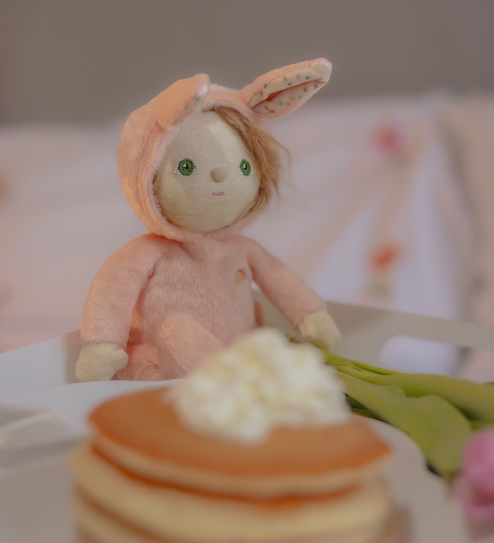 Olli Ella pink coloured Babbit Bunny Dinky Dinkum Doll in a seated position on a blanket. A pile of pancakes can be seen at the front. 