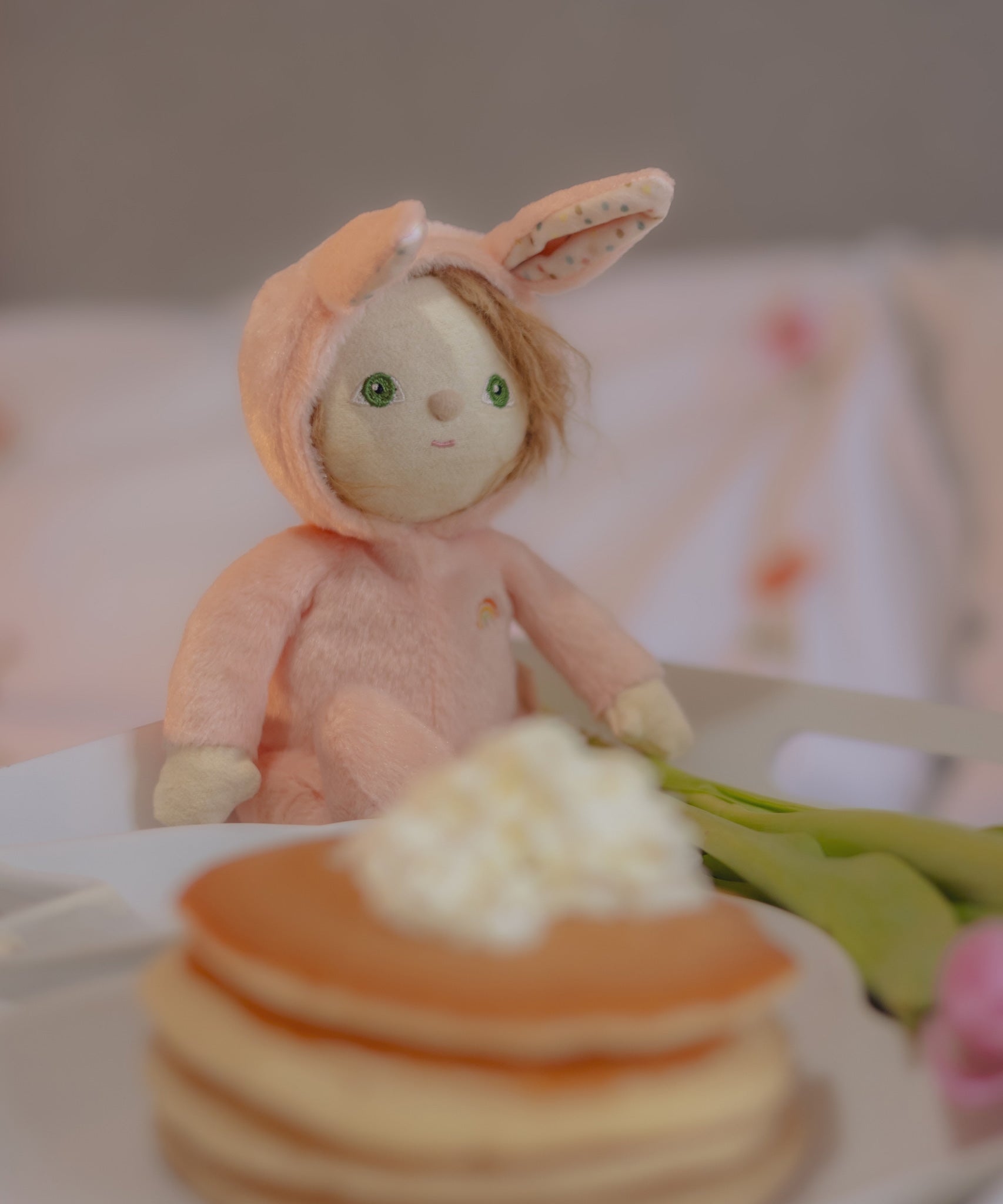 Olli Ella pink coloured Babbit Bunny Dinky Dinkum Doll in a seated position on a blanket. A pile of pancakes can be seen at the front. 