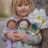 A child holding two Olli Ella Bunny Dinky Dinkum Dolls in their hands with a bunch of white flowers on their shoulder.