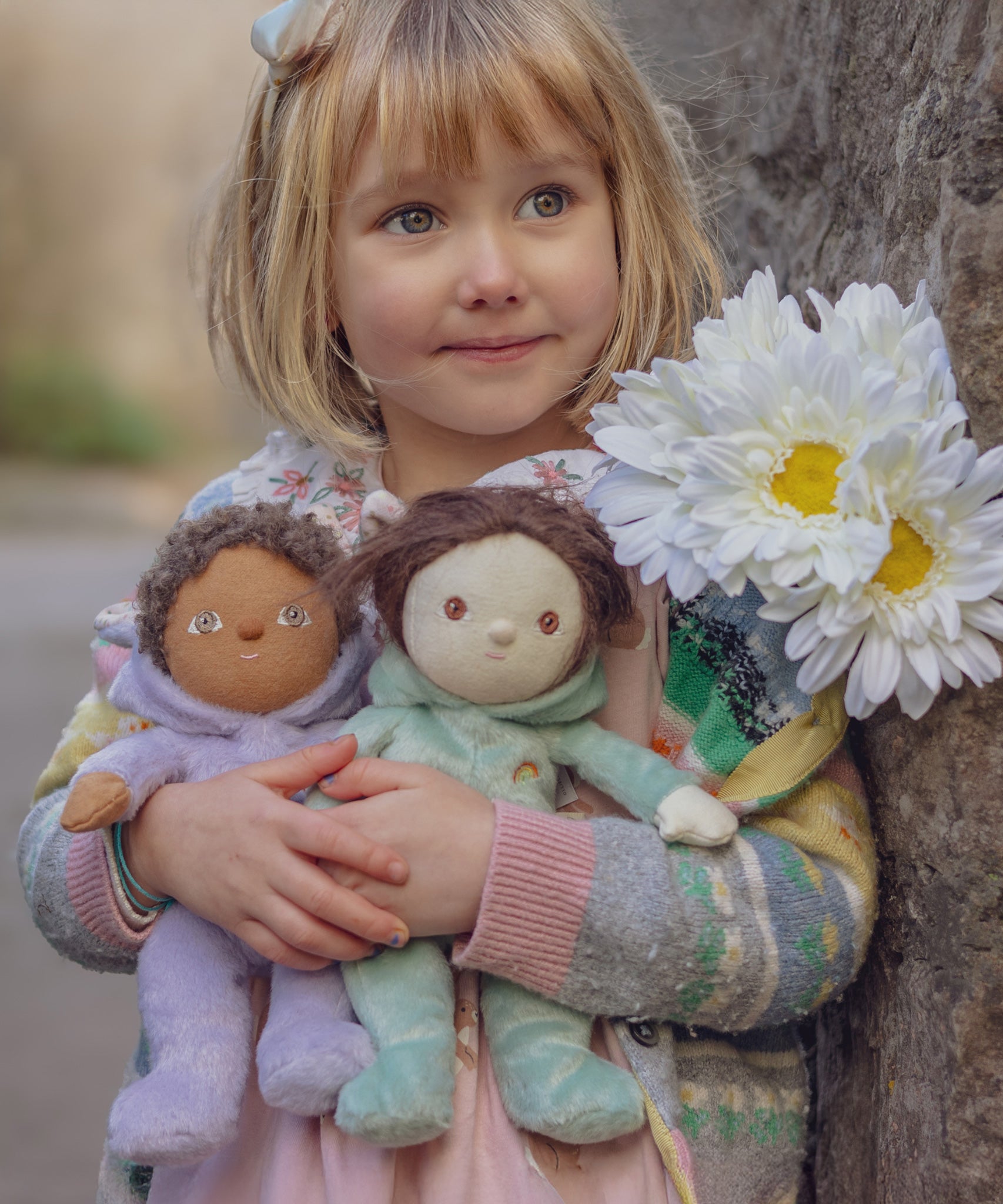 A child holding two Olli Ella Bunny Dinky Dinkum Dolls in their hands with a bunch of white flowers on their shoulder.