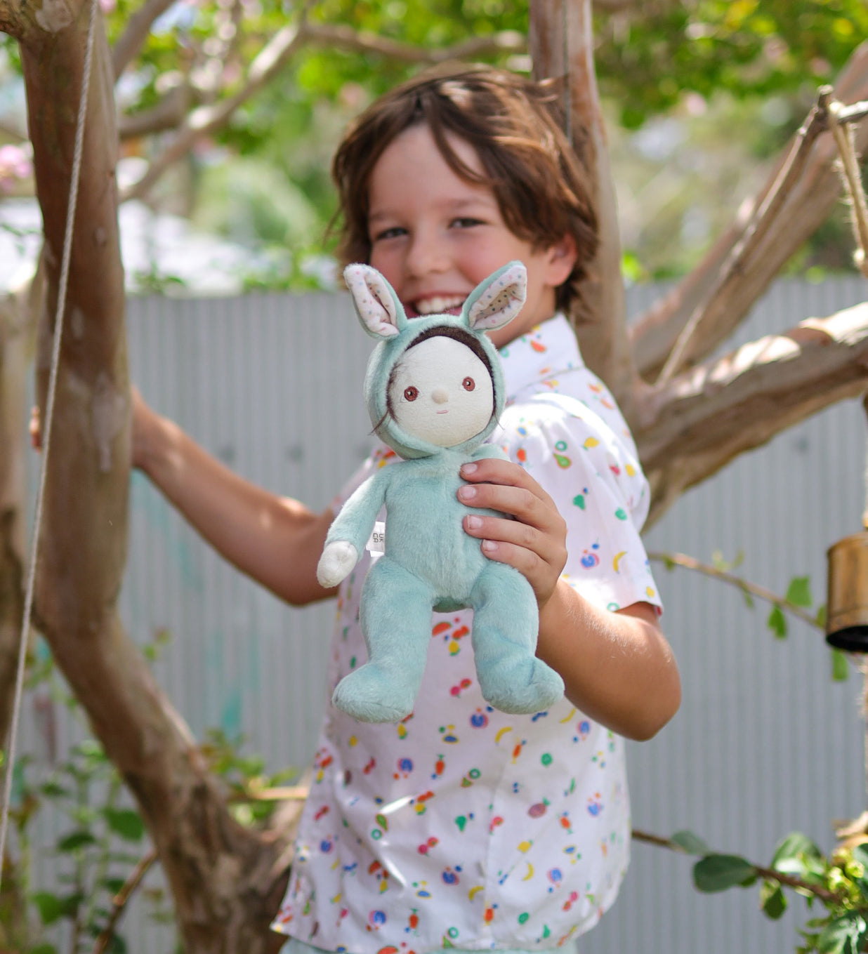 A child holding an Olli Ella mint green coloured Biscuit Bunny Dinky Dinkum Doll in their hand. They are standing under a tree.