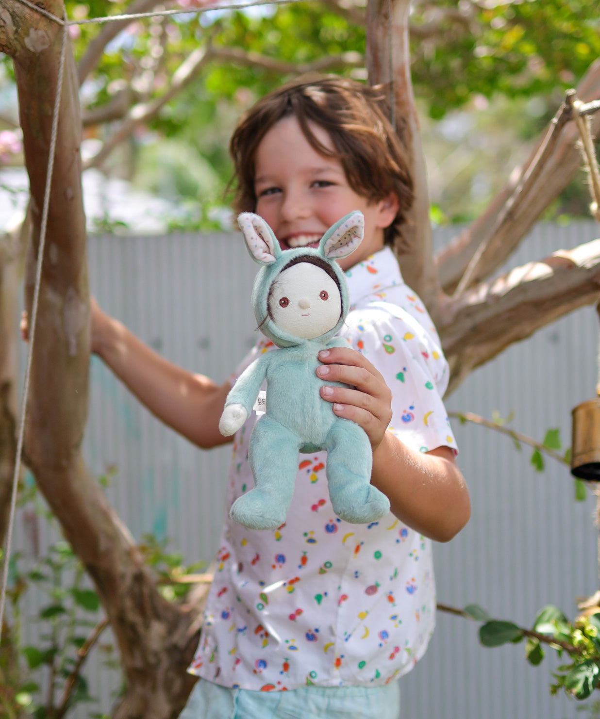 A child holding an Olli Ella mint green coloured Biscuit Bunny Dinky Dinkum Doll in their hand. They are standing under a tree.