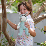 A child holding an Olli Ella mint green coloured Biscuit Bunny Dinky Dinkum Doll in their hand. They are standing under a tree.