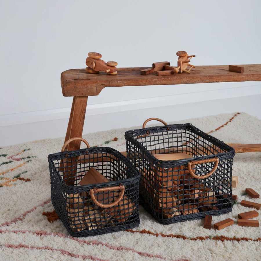 2 olli ella woven rattan cabouche baskets on a white carpet filled with wooden toys next to a wooden bench