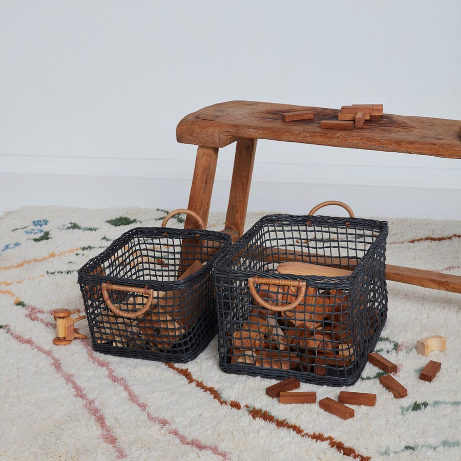 2 olli ella woven rattan cabouche baskets on a white carpet filled with wooden toys next to a wooden bench