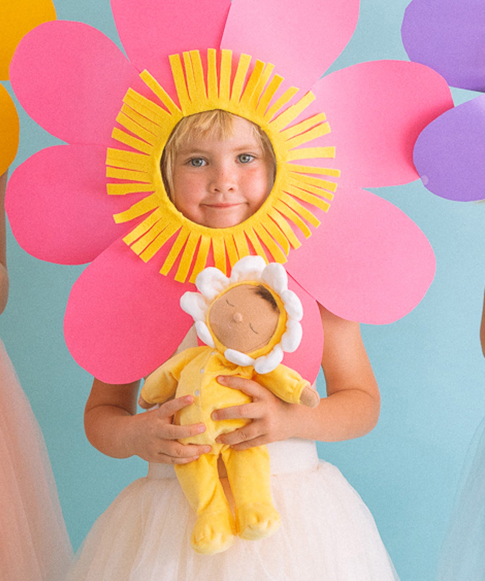 A child wearing a big pink flower headdress holding on to a Olli Ella Buttercup Pip Dozy Dinkum Doll.